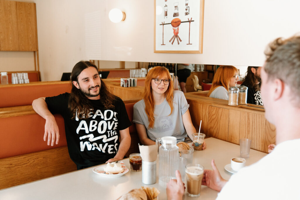 A group of men and women sit in a cafe wearing Heads Above The Waves merch, talking about mental health and drinking coffee. 