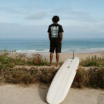 A man stands facing the ocean, wearing a black t-shirt from Heads Above The Waves and Reef. A white surfboard is next to him.