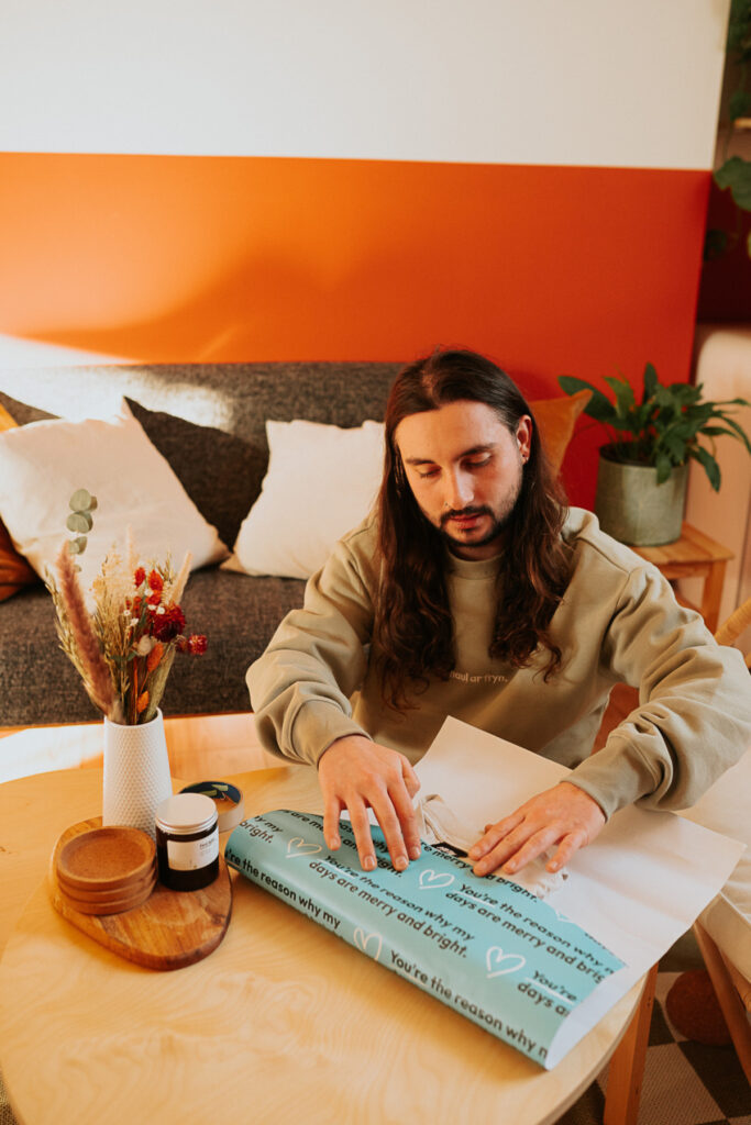 A man with long hair is wrapping a present with wrapping paper that says "you're the reason why my days are merry and bright" 